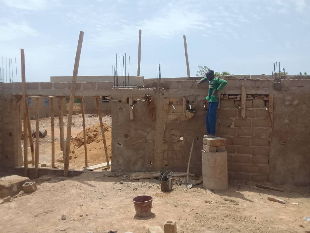 Un maçon en équilibre instable sur des briques de ciment sur un chantier informel à Bamako, illustrant le manque d'équipements de sécurité et l'amateurisme dans le BTP au Mali.